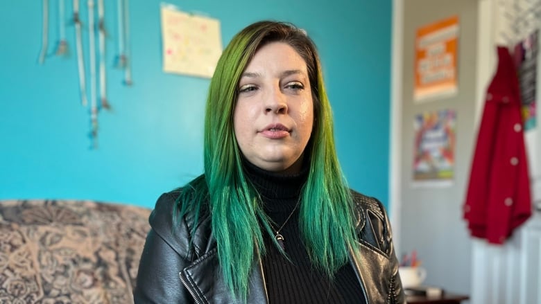 Gabrielle Giroux, with hair dyed green, sits on a couch in front of a bright teal wall. 