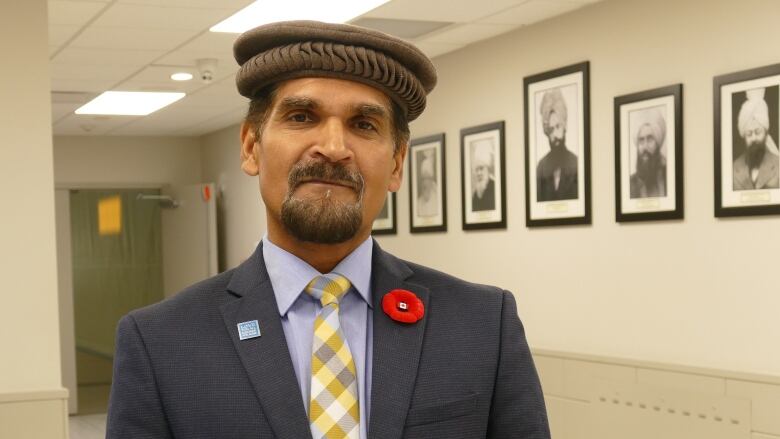 A man stands in a hallway. On a wall behind him, there hangs six portraits of various Muslim men. The man is wearing a charcoal grey suit, a yellow plaid tie, a light blue shirt and a brown hat. A poppy, with a mini Canada flag pin, is on his lapel.