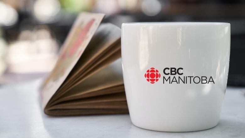 A white coffee mug with CBC Manitoba branding next to a book on a table. 