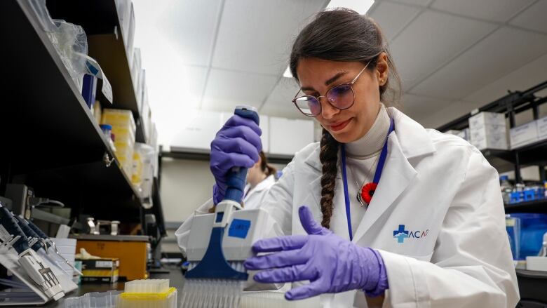 A woman in a white lab coat and purple gloves handles samples on a counter in a lab.