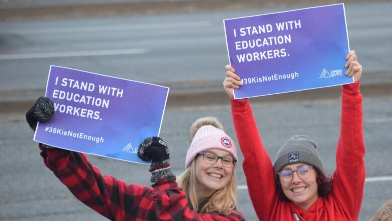 Two young women wearing toques hold up signs reading 'I stand with education workers' during a protest along the Kingsway in Sudbury