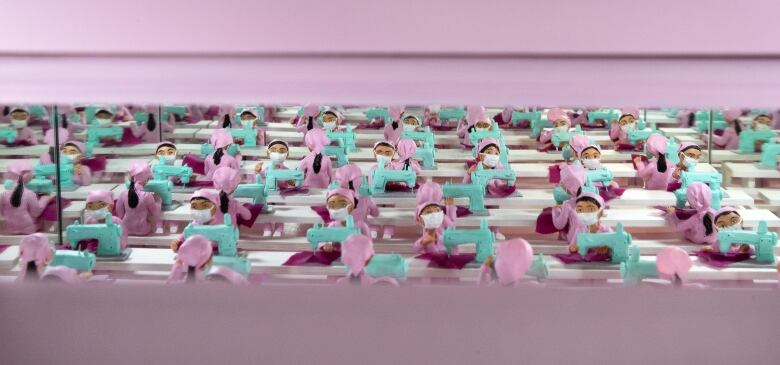 Diorama of miniature figurines. The figures are garment factory workers, seated at seemingly infinite rows of sewing machines. They wear face masks and pink smocks and kerchiefs.