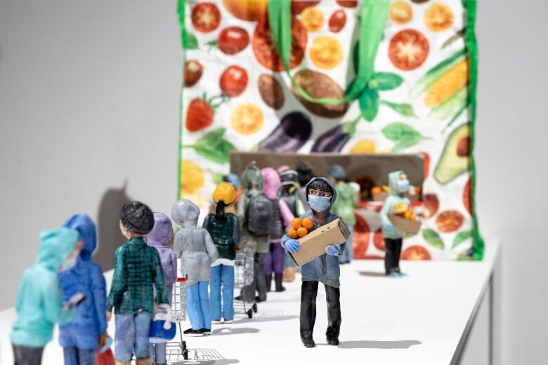 Mini figurines of people are lined up on a white countertop to access a printed reusable grocery bag. The figures push empty shopping carts and wear winter clothing and face masks.