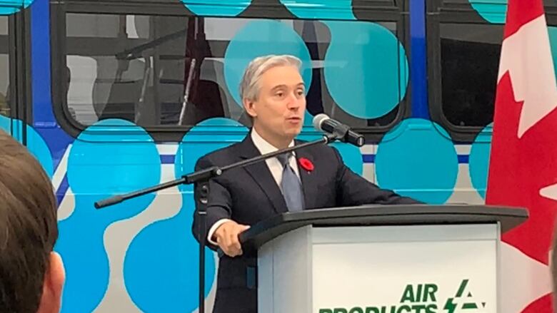 A man with white hair and wearing a dark suit and tie speaks at a podium.