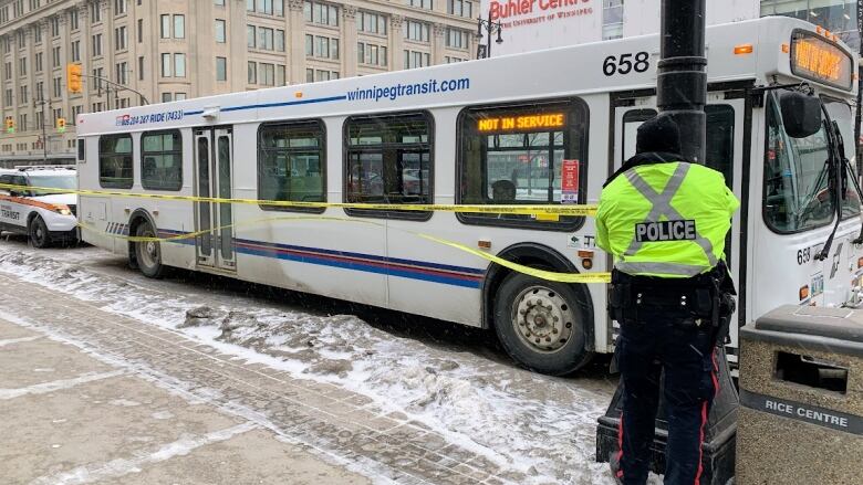 White bus on the street in front of the Buhler Centre on Portage Avenue, with a police officer securing yellow police tape to a lamp post beside it.