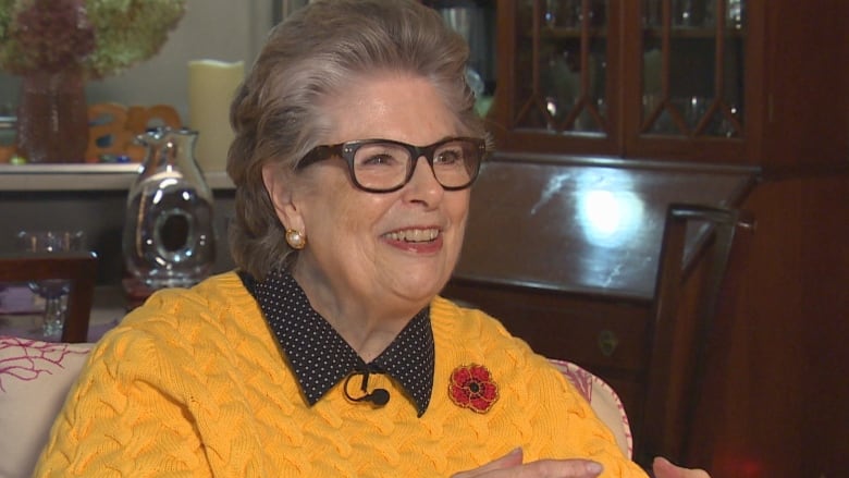 Close up of a smiling woman with brown hair and glasses, wearing a yellow sweater, wearing a Remembrance Day red poppy pin to honour veterans.