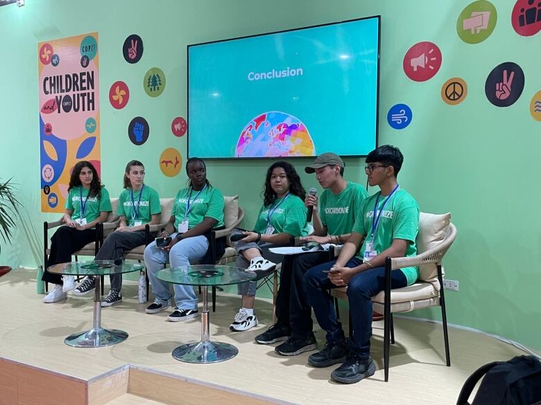 Several youths are sitting around a short glass table, in front of a green wall with a television hanging on it. Each of the youths are wearing a green t-shirt. One is speaking into a microphone.