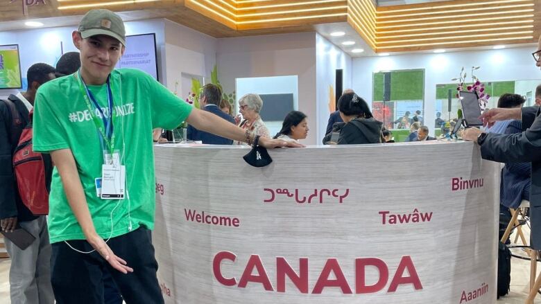 A young man wearing a green t-shirt and a beige ball cap is posing next to a podium. The podium, which is made of grey laminated wood, reads 'Canada' and has the word 'Welcome' in various languages pepperred throughout. The words are coloured red.