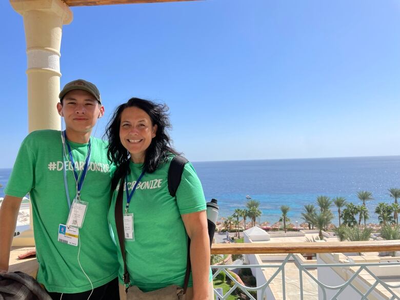 A young man and a woman stand next to each other, smiling. They're each wearing green t-shirts that read, 'hashtag Decarbonize,' and accreditation passes. The man is wearing a beige ball cap. The woman is wearing a knapsack, with a water bottle sticking out of the side. They are standing on a balcony. Behind them is wide blue ocean and a clear blue sky. There are a few palm trees in the background too.