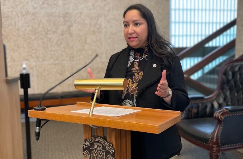 A woman with long black hair, wearing a black pant suit and brown, white and black printed shirt, stands behind a wooden podium with a gold metal desk lamp in front of her.