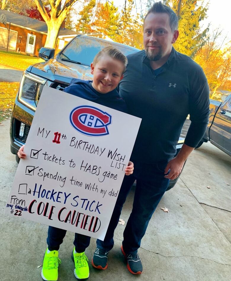 A father and son stand together outdoors. The son's sign says 