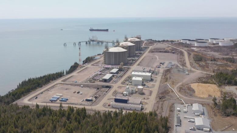 An aerial view of an industrial facility along a shoreline, featuring three large metal cylinders used for storage of liquefied natural gas. 