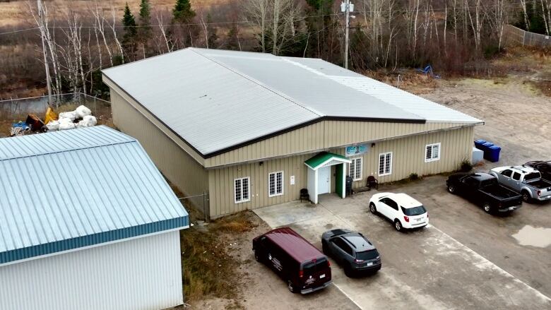 A square building is shown from above. It has a silver metal roof and brown siding. There are barred windows on the front and a large wooded area behind. 