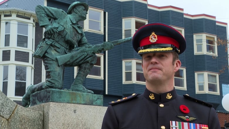 A man wearing a military uniform stands in front of a statue of a soldier. He has several military medals on his uniform, and is wearing a poppy above them.