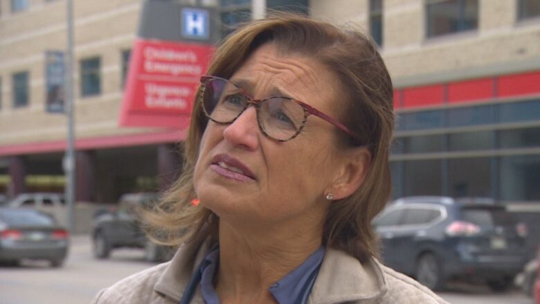 A woman with glasses and a stethoscope around her neck looks distressed outside a hospital.