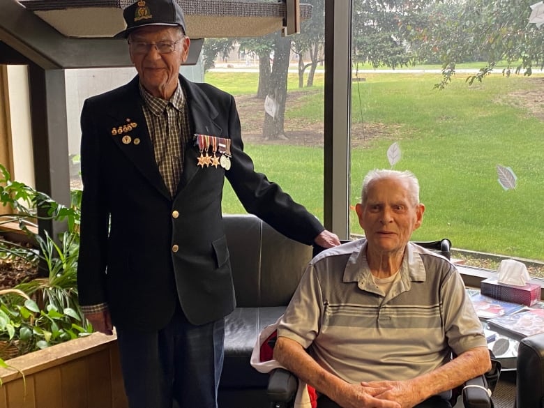 A an elderly man decorated in military medals stands beside another elderly man who sits in a chair. 