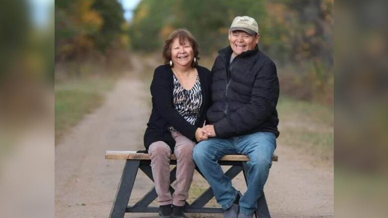 An older couple sits on a bunch on a rural road holding hands and smiling.