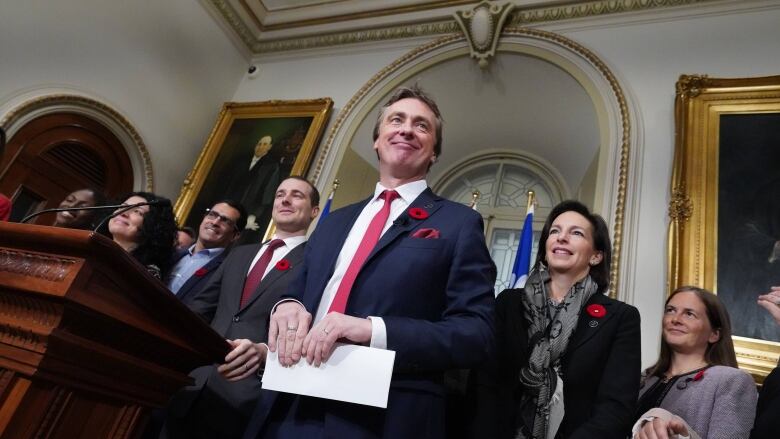 An angled shot of a guy in a suit, surrounded by men and women with an ornate background.