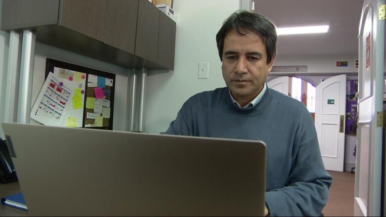 A man sits clad in a blue grey sweater working on his laptop.