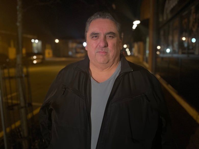 A man stares straight at the camera. The sky is dark.  The man is wearing a black coat and a grey t-shirt.