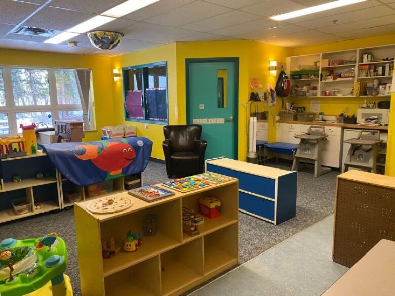 A children's playroom filled with shelves and toys, high chairs and a kitchenette. The walls are painted a bright yellow and everything is neatly organized.