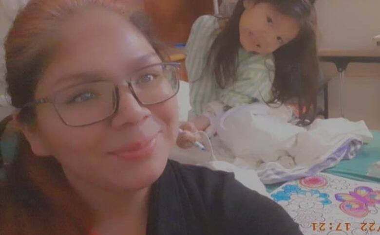 A woman with glasses smiles in a selfie as a young girl poses and smiles on a hospital bed covered with colouring supplies behind her.