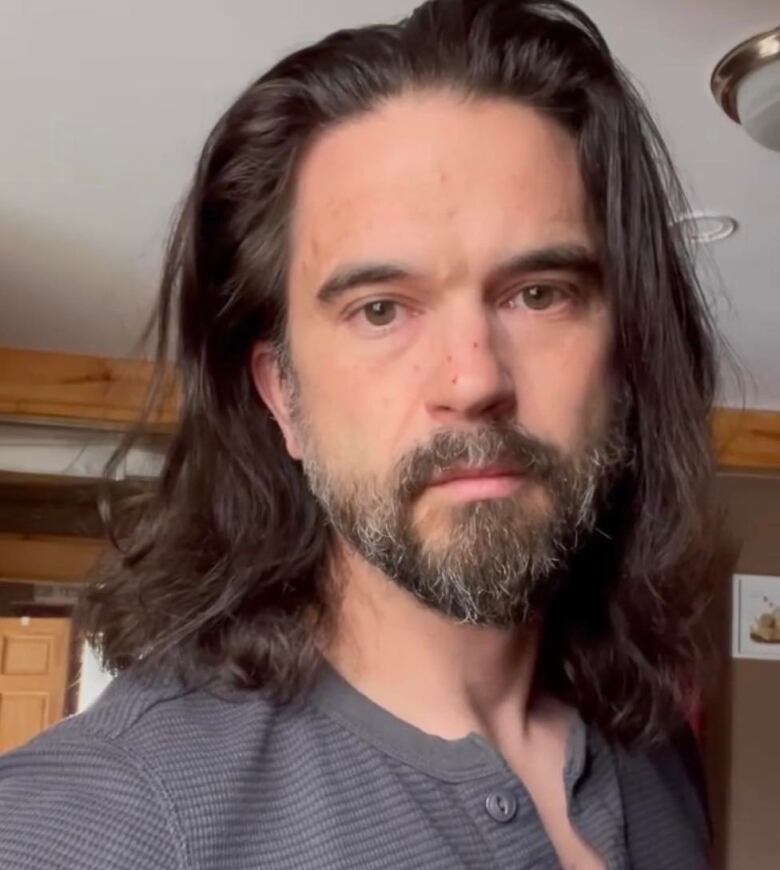 A close-up view of a man's face, with long hair, beard, and mustache, looking directly into the camera.