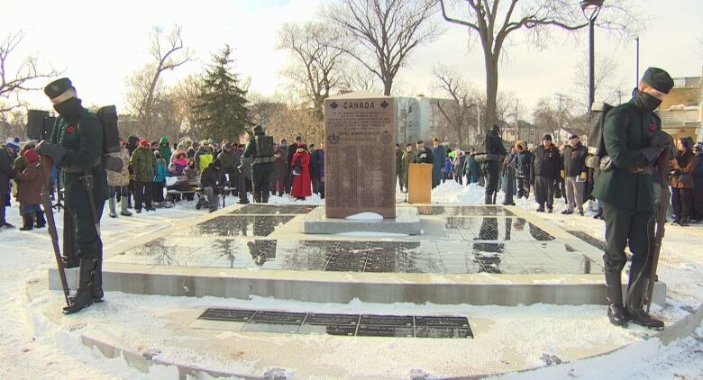 A crowd of people stand in snow, gathered around a cenotaph and soldiers.