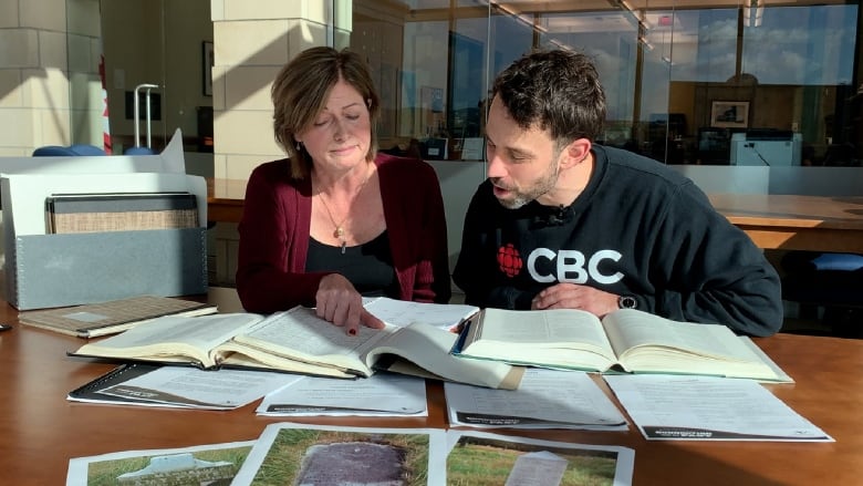 A woman and a man sit side by side at a table, crowded with books and records. The woman points to a particular record as the man reacts with surprise.