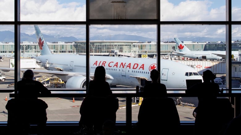 A row of people, in silhouette, wait at an airport departure lounge with an Air Canada plane visible in the background.