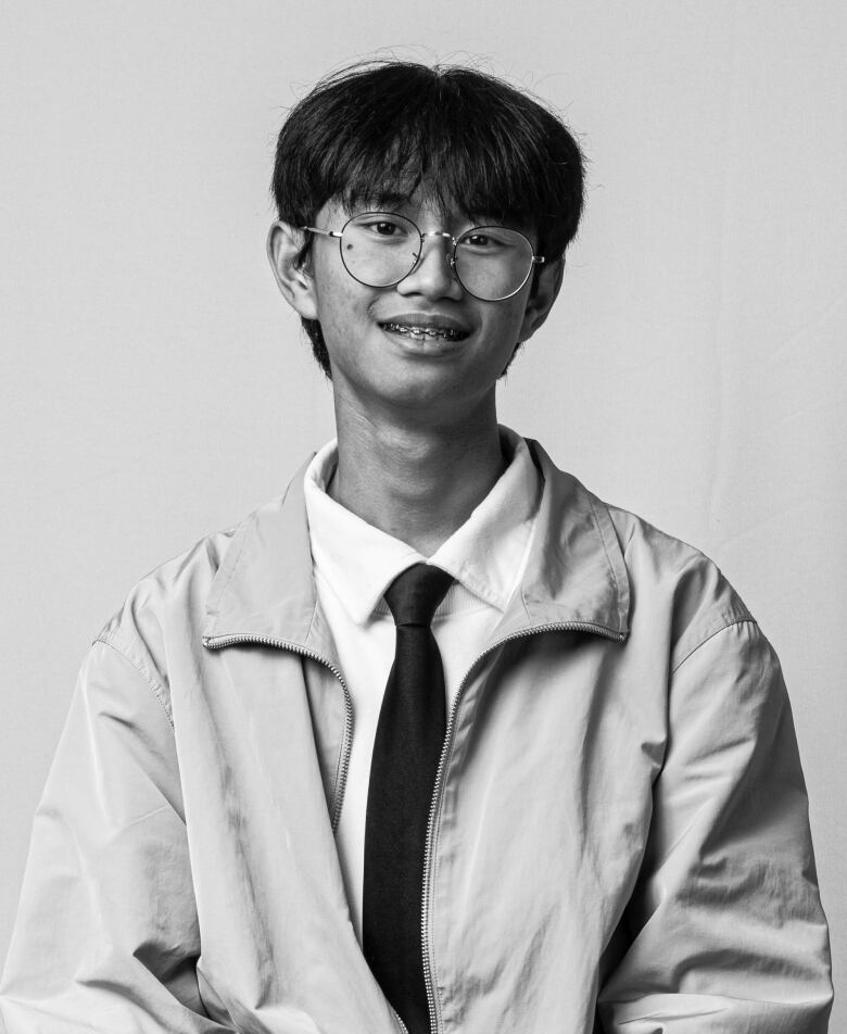 A young man wearing glasses, a shirt and tie and a zippered jacket smiles at the camera in a black-and-white photo.