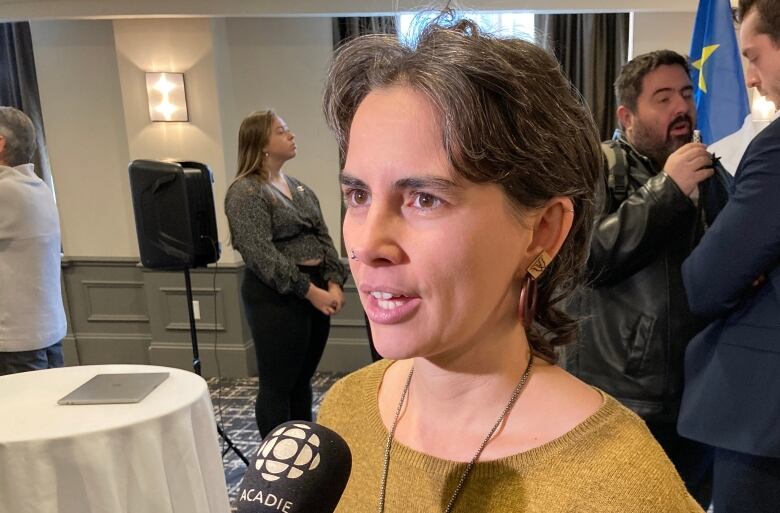 A woman speaking into a mic with the CBC gem on it. She is wearing a mustard yellow shirt. Two men are talking in the background. 