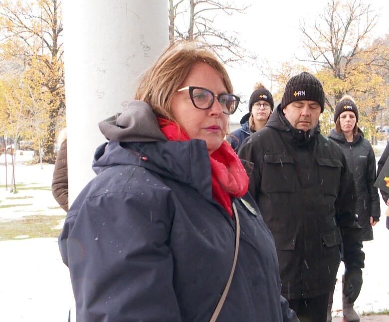 A woman wearing a navy blue jacket with glasses and a red scarf stands with a group of people inside a gazebo.