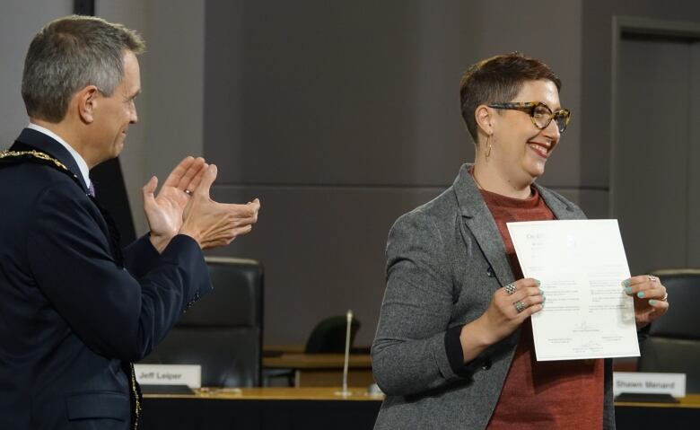 A woman holds a document as a man looks on, clapping.