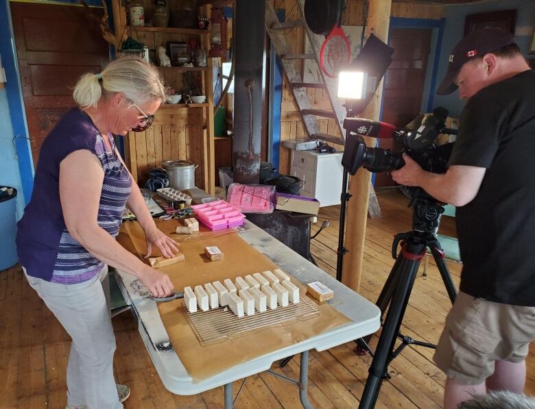 A standing woman cuts bars of soap on a work table. To her right, a man uses a video camera on a tripod to record the scene. 