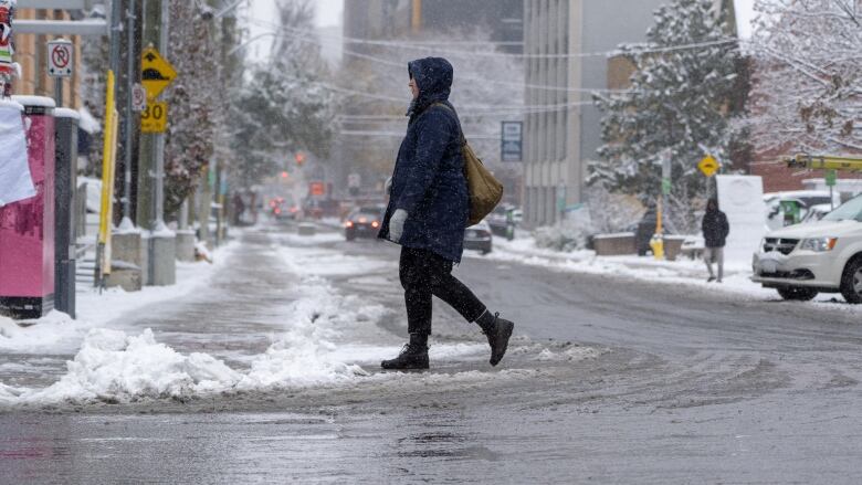 Someone in a winter coat, boots and mittens crosses a street as it snows.