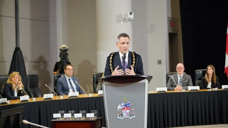 A man speaks in front of a podium while several people sit behind him.