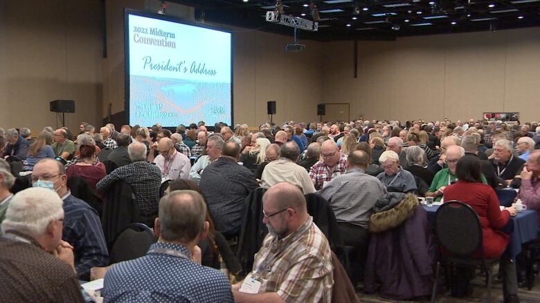 hundreds of people sat at tables with a large projected monitor behind them