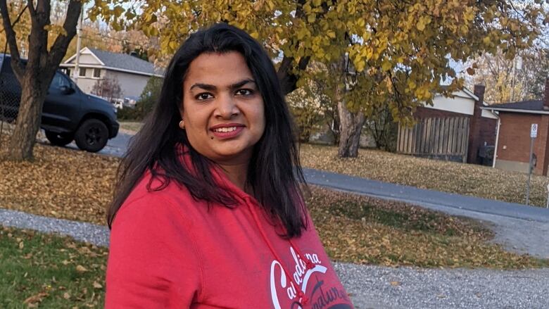 Anjali Aggarwal stands in a driveway wearing a pink sweater, with autumn leaves on the ground behind her. 