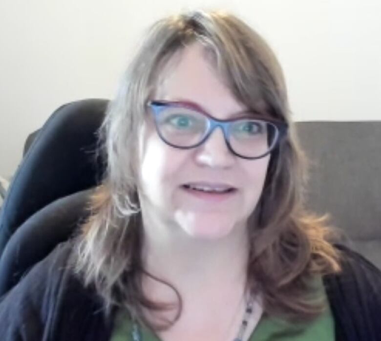 A woman with brain hair and glasses sits in front of a couch.