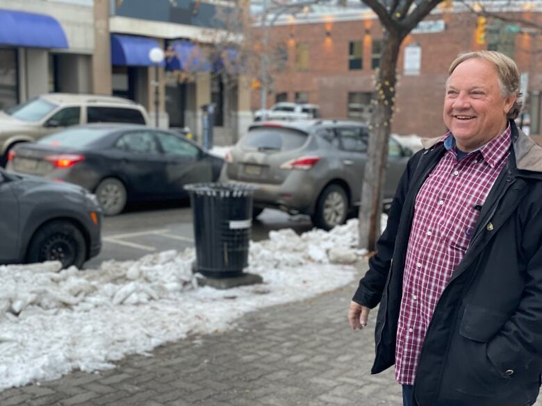 A middle-aged man in a red and white plaid shirt and black winter coat stands smiling on a sidewalk, with a small bank of dirty snow behind him.