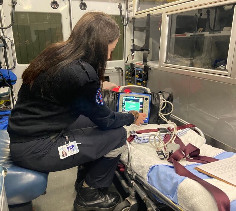 A female paramedic sits inside an ambulance with an automated external defibrillator layed on top of a stretcher. She is pressing a button on the defibrillators. 