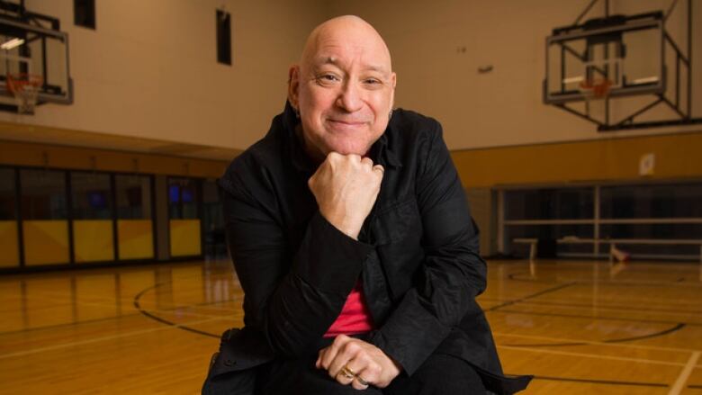 A bald man sits in a wheelchair in the centre of a gymnasium and stares into the camera.