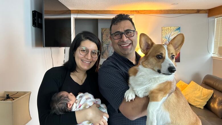 A man stands holding his corgi dog. A woman stands next to his left with a baby in her hands. They are smiling.