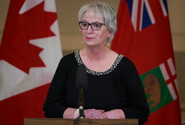 Eileen Clarke stands at a podium with the flags of Canada and Manitoba behind her.