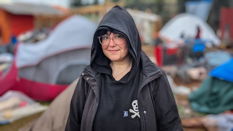 A woman with glasses, a black hoodie and a black jacket smiles at the camera. She is standing outside and there are tents in the background. 