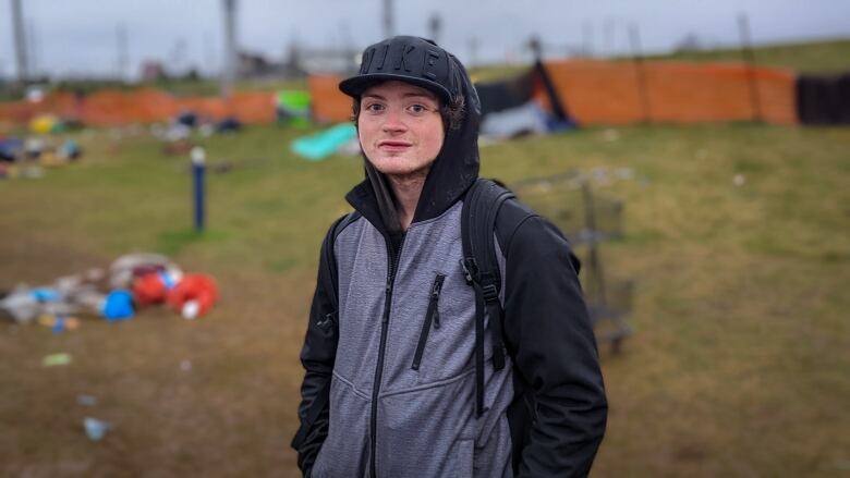 A young man wearing a black and grey jacket, a black ball cap and a backpack looks at the camera. He is standing outside and there are tents in the background. 