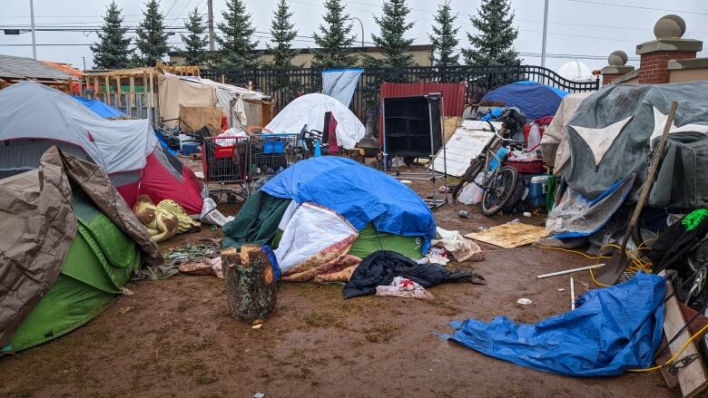 Several tents are set up close together on muddy ground. Some have blankets or tarps thrown on top of them. We also see a bicycle, coolers, and other items on the ground near the tents. 