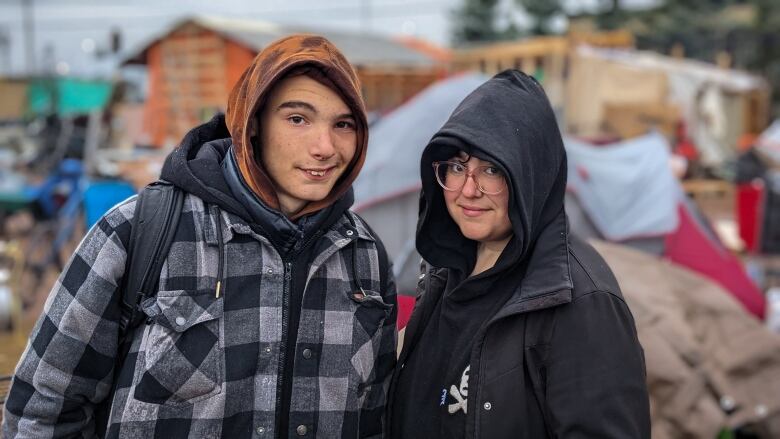 A young man and woman look at the camera. The young man is wearing a plaid jacket and a brown hoodie. The woman is wearing a black jacket and a black hoodie. They are standing outside and there are tents behind them. 