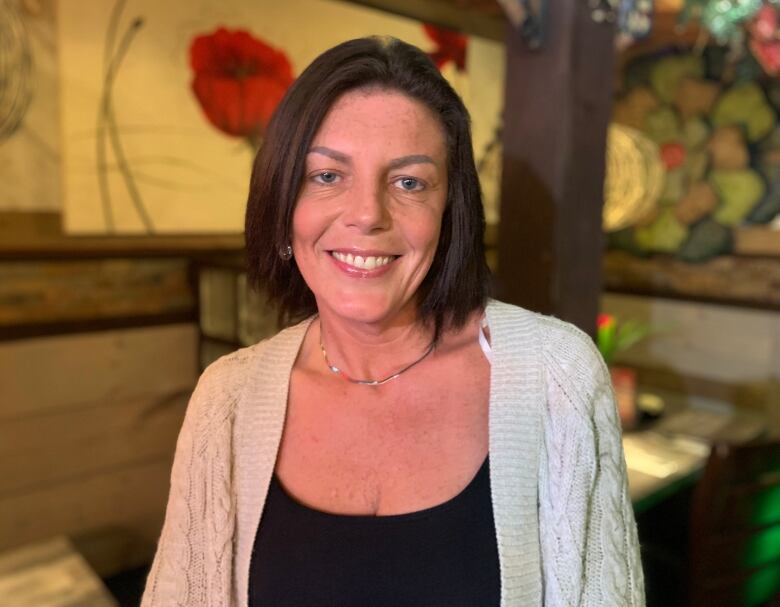 A head and shoulders shot of a person with shoulder-length hair stands in front of a some tables in a restaurant.